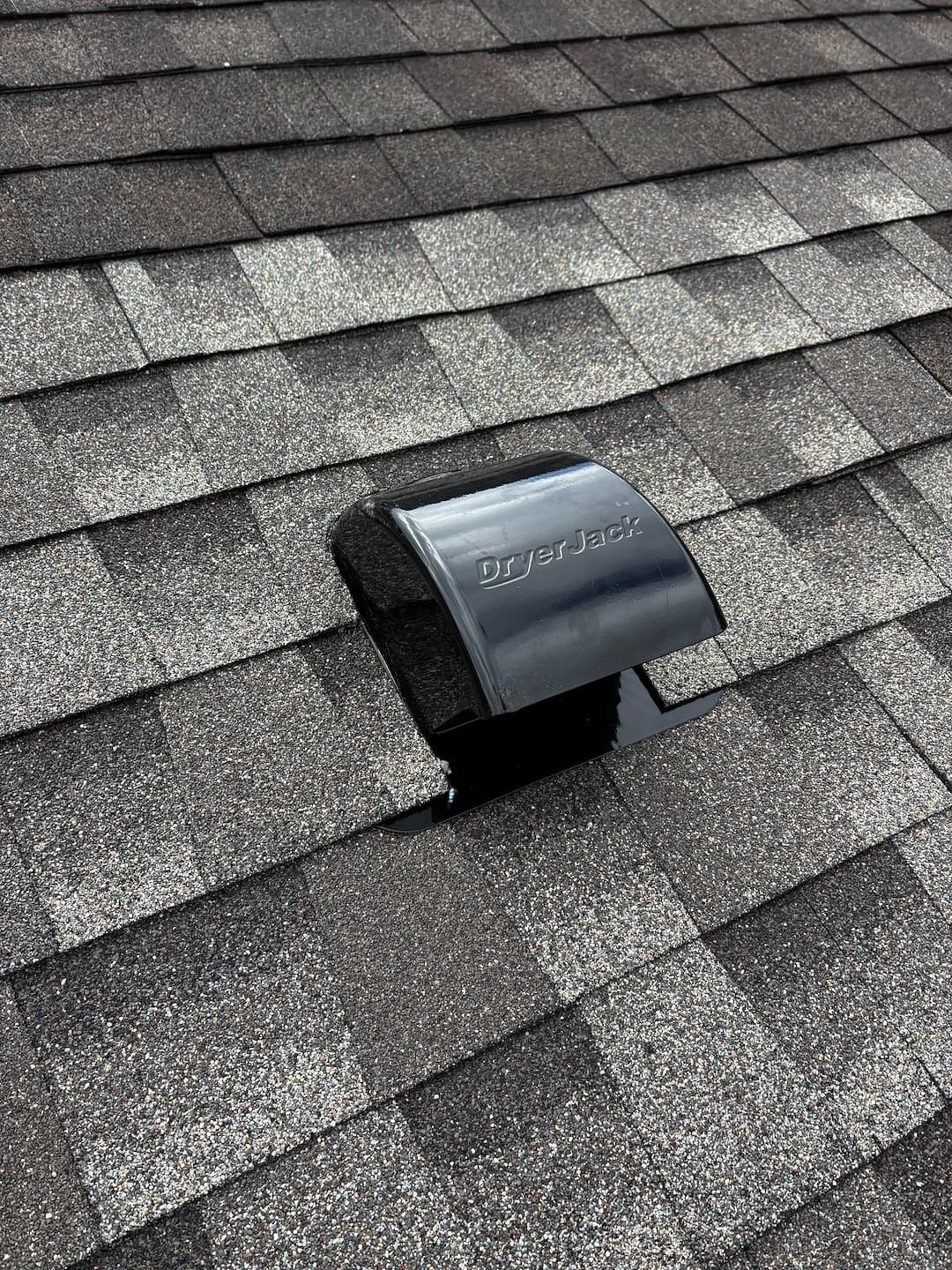 Black DryerJack roof vent cap installed on a sloped roof with grey asphalt shingles.
