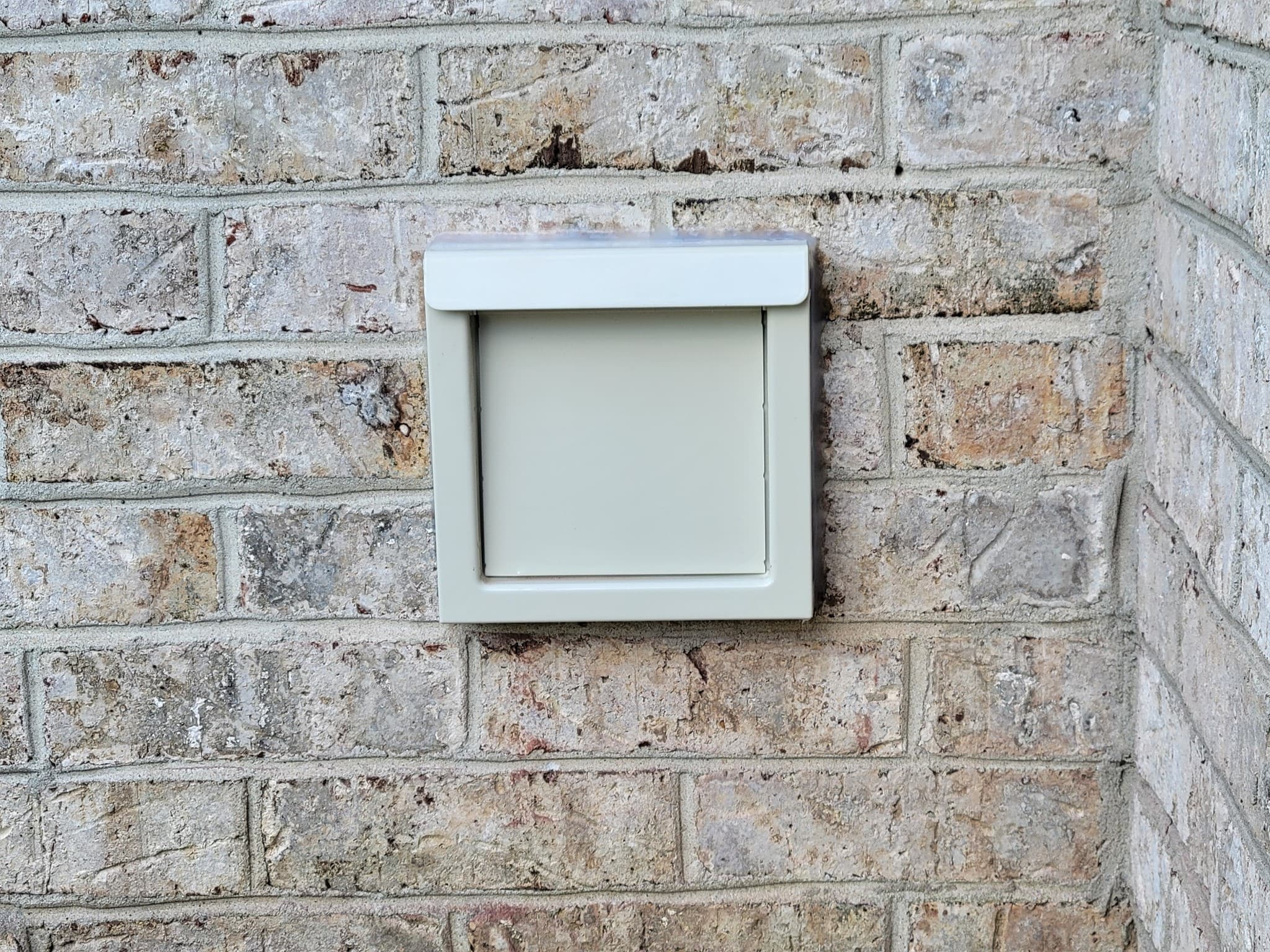 Square beige plastic vent cover with a flap mounted on a light brick wall.