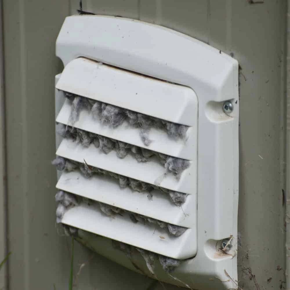 White exterior dryer vent cover heavily clogged with thick grey lint between the horizontal slats.